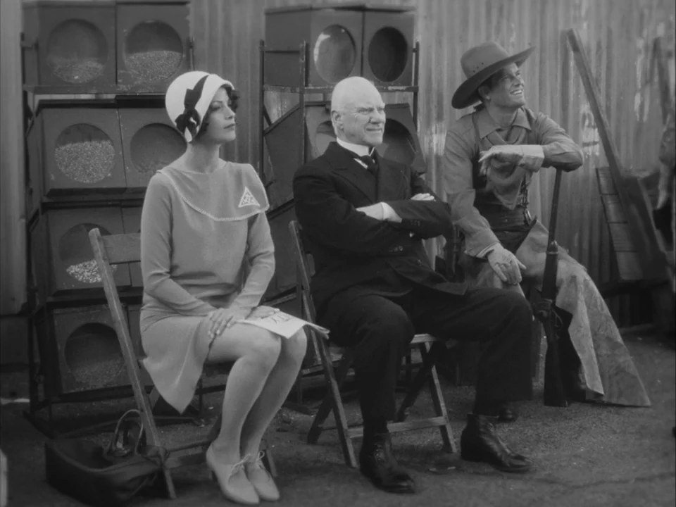 The Artist (2011) movie still: fullbody — Three people sit in folding chairs in front of a corrugated metal wall. They are positioned in a row and…