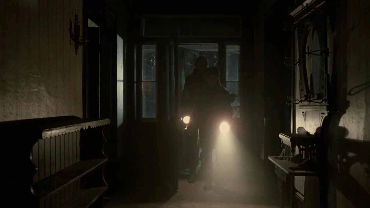 Splice (2009) movie still: medium — A dimly lit interior hallway with dark walls and an antique wooden wall unit. Two figures enter from an open…