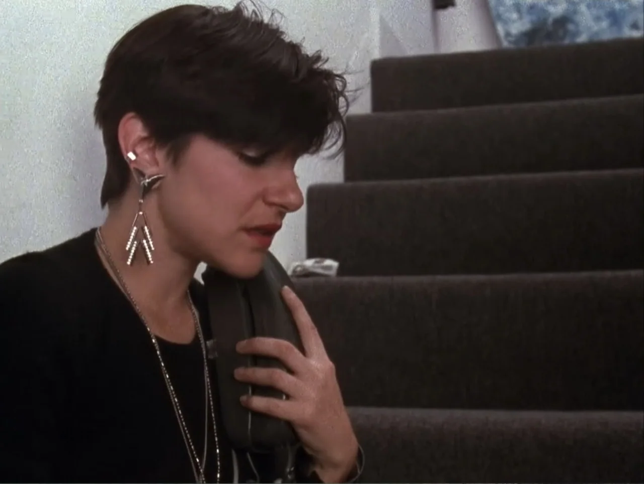 The Living End (1992) movie still: closeup — A dark carpeted staircase provides the backdrop for a person standing against a wall. A young woman…