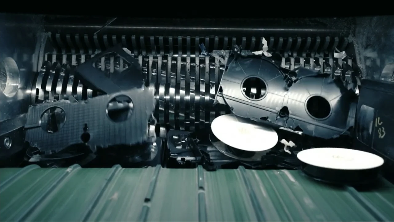 The Forever Prisoner (2021) movie still: closeup — A close-up view of a heavy-duty industrial shredder containing black plastic cassette tape…
