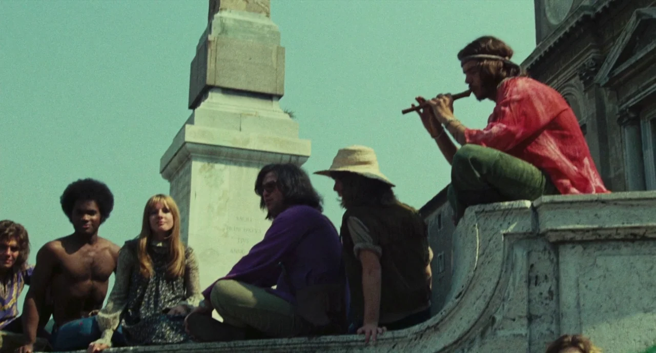 Roma (1972) movie still: fullbody — A group of young adults sits on stone steps near an obelisk. One man plays a wooden flute while others look on…