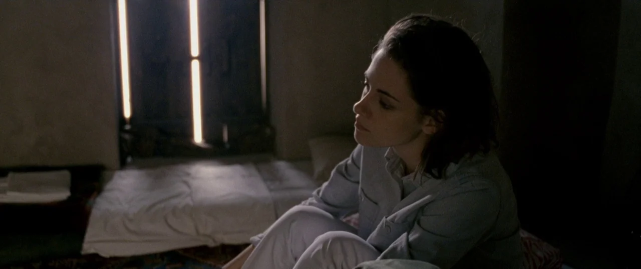 Personal Shopper (2016) movie still: medium — A dimly lit, sparse room with concrete walls and a floor-level bed frame. A woman sits on the bed,…