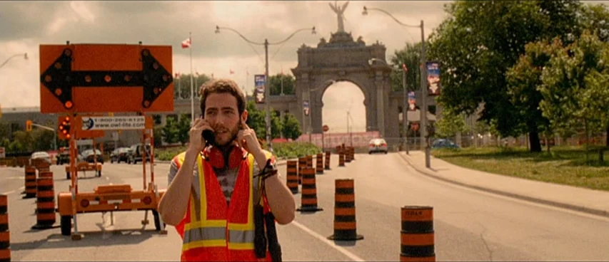 This Movie Is Broken (2010) movie still: medium — A man in a safety vest stands on an urban road with traffic cones. In the background stands a…