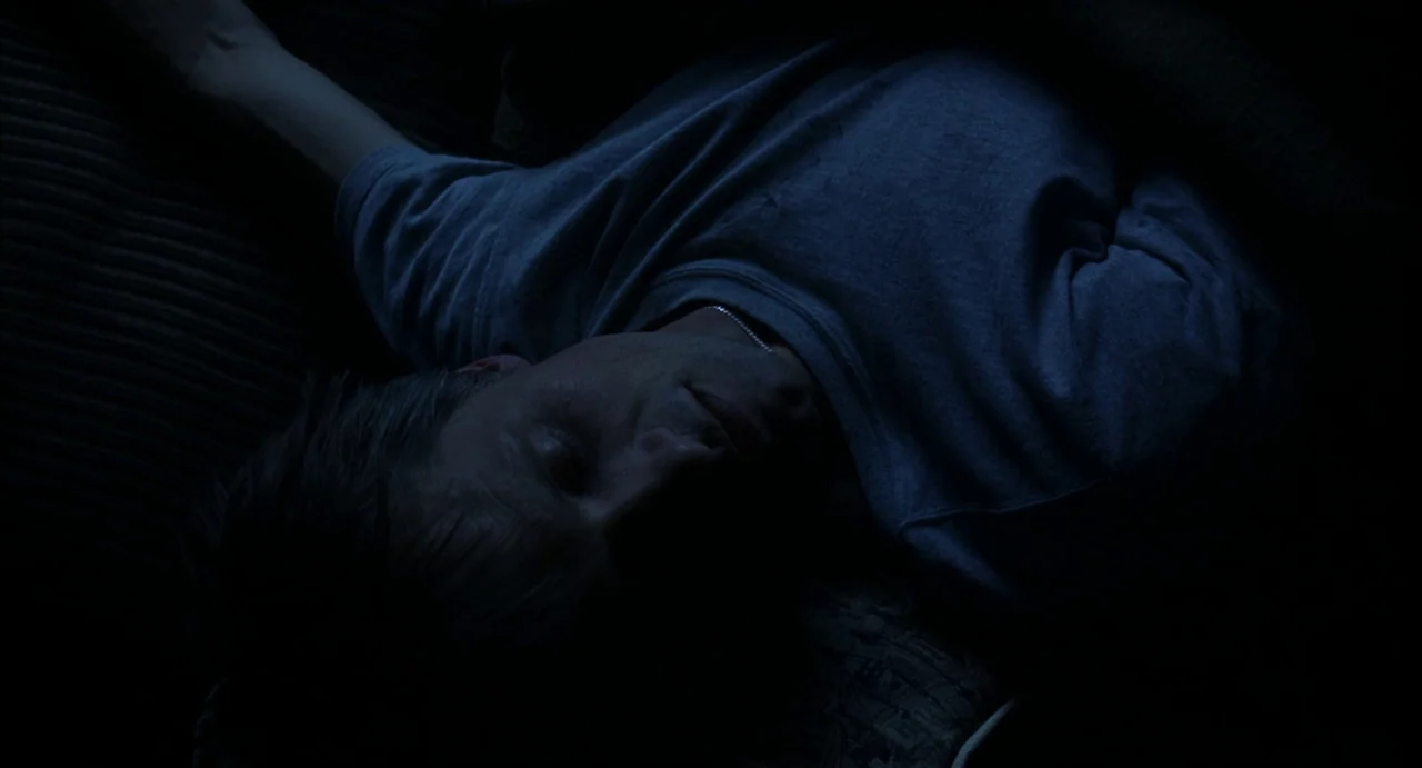 A History of Violence (2005) movie still: closeup — A man sleeps on a couch in a dark, blue-lit room. He lies with eyes closed while wearing a…