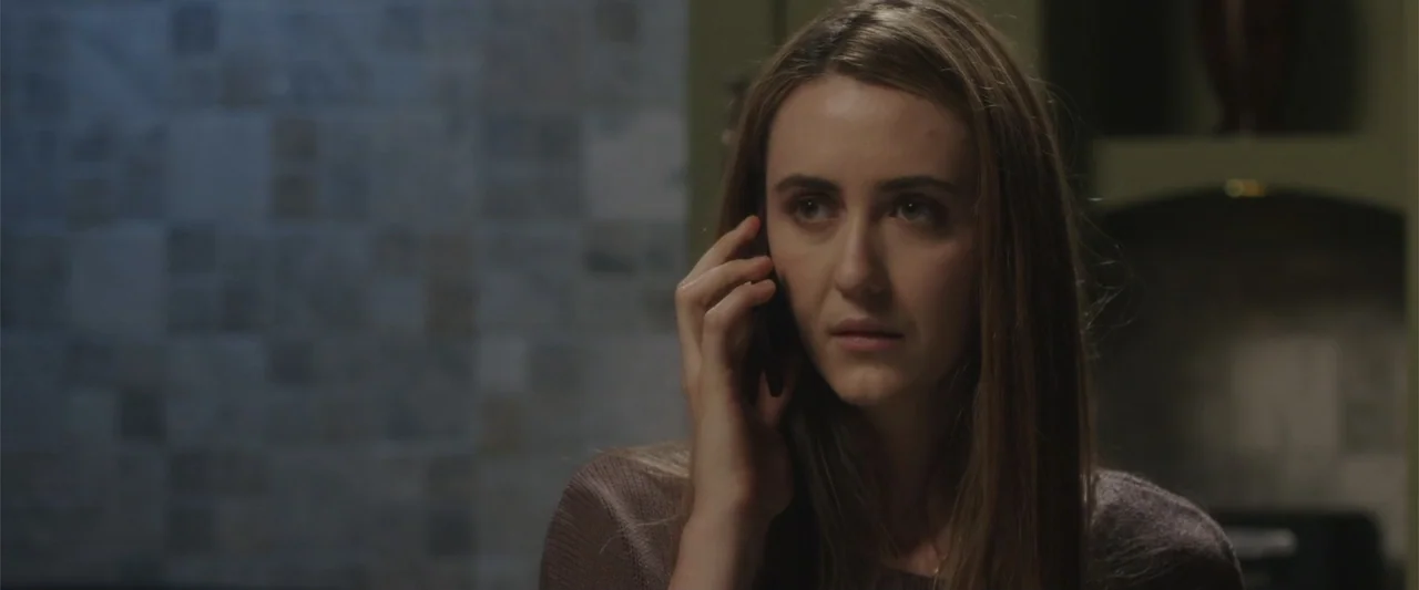 Presagios (2024) movie still: closeup — A dimly lit interior space features a wall with neutral-colored square tiles. A young woman with long brown…