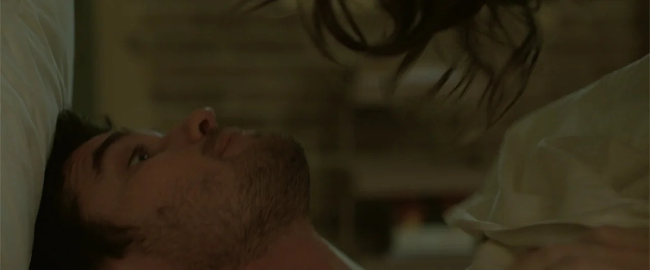 Marked Men: Rule + Shaw (2025) movie still: closeup — A dimly lit bedroom features a man lying on a white pillow. A second person is partially…