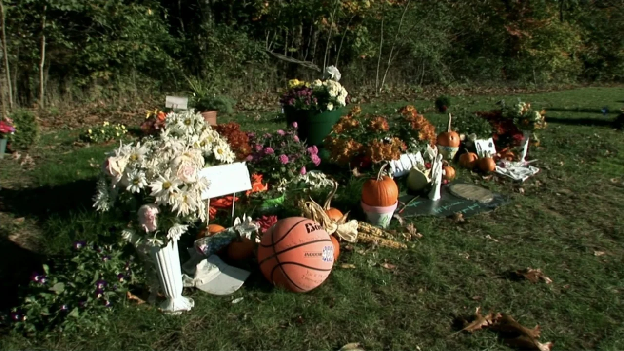 The Cheshire Murders (2013) movie still: medium — A grassy burial plot is covered in flowers, pumpkins, and a basketball. The setting is a bright…