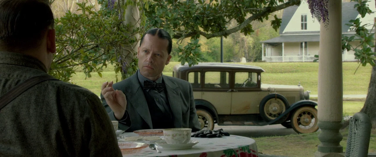 Lawless (2012) movie still: medium — A wooden porch with a table set for tea overlooks a rural residential property. A man in a suit sits at the…