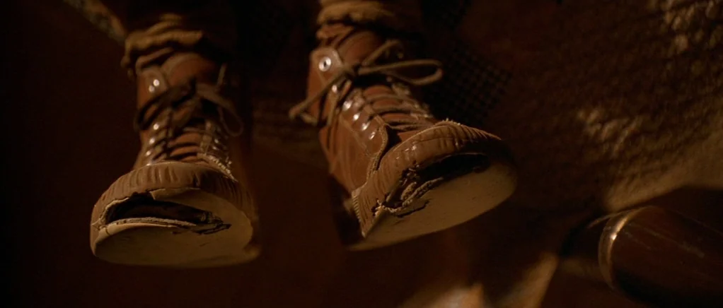 King of the Hill (1993) movie still: closeup — A pair of worn brown leather sneakers with torn soles hang in a dark space. The shoes are suspended…