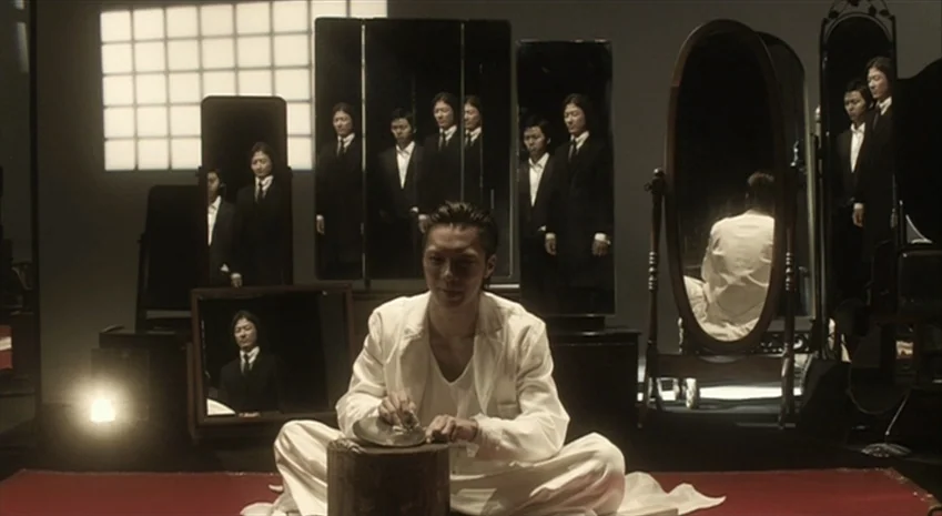 Rampo Noir (2005) movie still: medium — A spacious room contains many tall mirrors reflecting men in black suits. A man in white sits centrally on a…