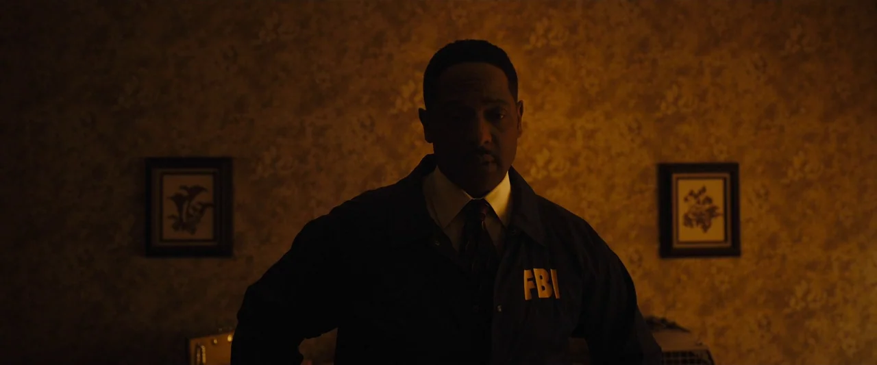 Longlegs (2024) movie still: medium — A man stands in a dimly lit room with vintage patterned wallpaper. Two framed pictures hang on the wall behind…