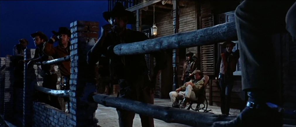 Fistful Of Dollars (1964) movie still: wide — A dark wooden porch at night with several men standing behind a wooden railing. One man sits in a…