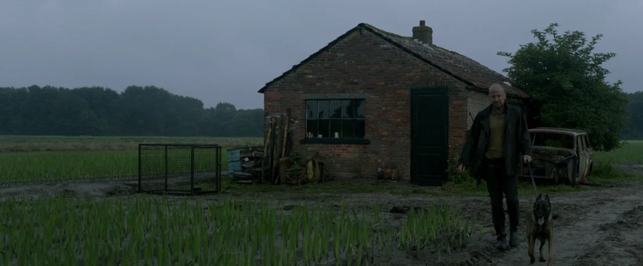 Borgman (2013) movie still: wide — A red brick shed stands beside a muddy field under a heavy gray sky. A man walks along the path with a…
