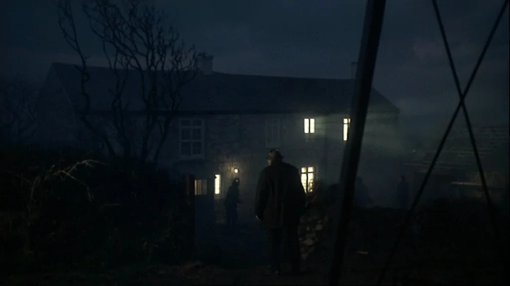 Straw Dogs (1971) movie still: wide — A stone farmhouse sits in a dark, foggy field during the night. A man stands in the foreground looking towards…