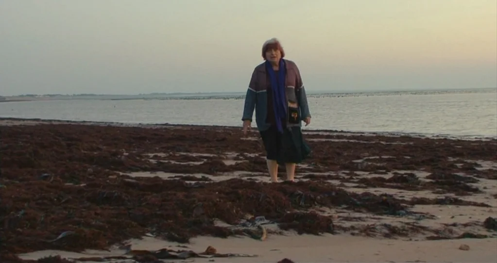 The Beaches of Agnès (2008) movie still: fullbody — An elderly woman stands on a desolate beach covered in mounds of dark seaweed. The calm water…