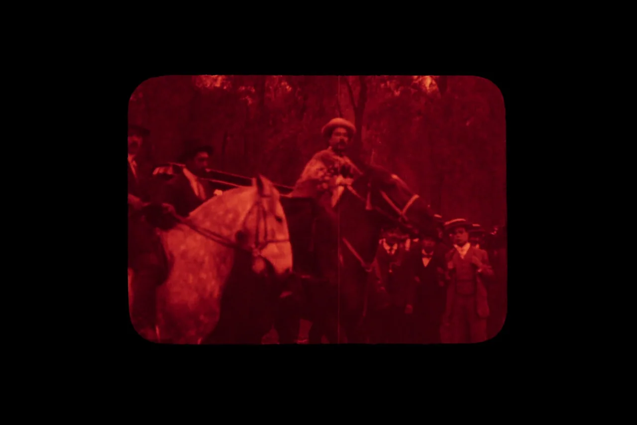 The Settlers (2023) movie still: wide — A group of men on horses move through a dense, tree-filled outdoor setting. The footage is tinted entirely…
