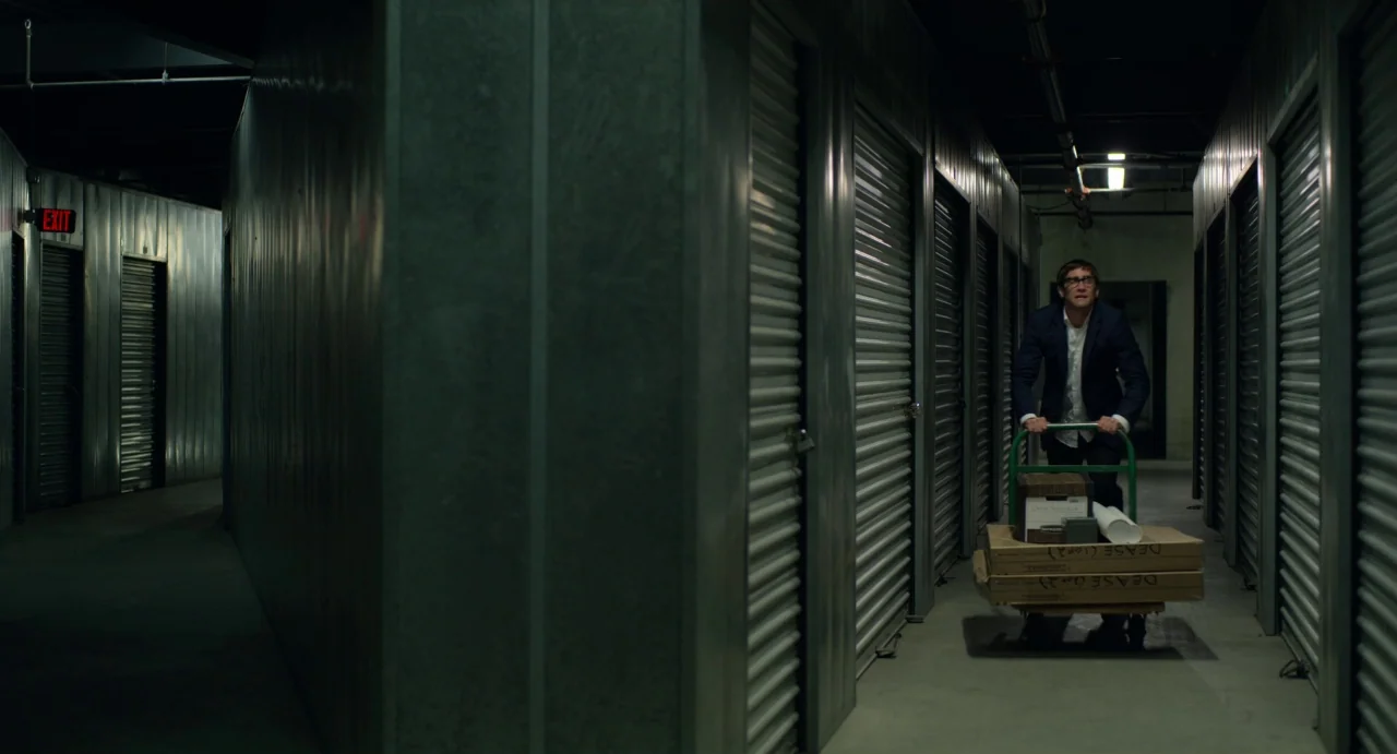 Velvet Buzzsaw (2019) movie still: wide — A narrow corridor features rows of metal roll-up doors on both sides. A man wearing a suit pushes a cart…
