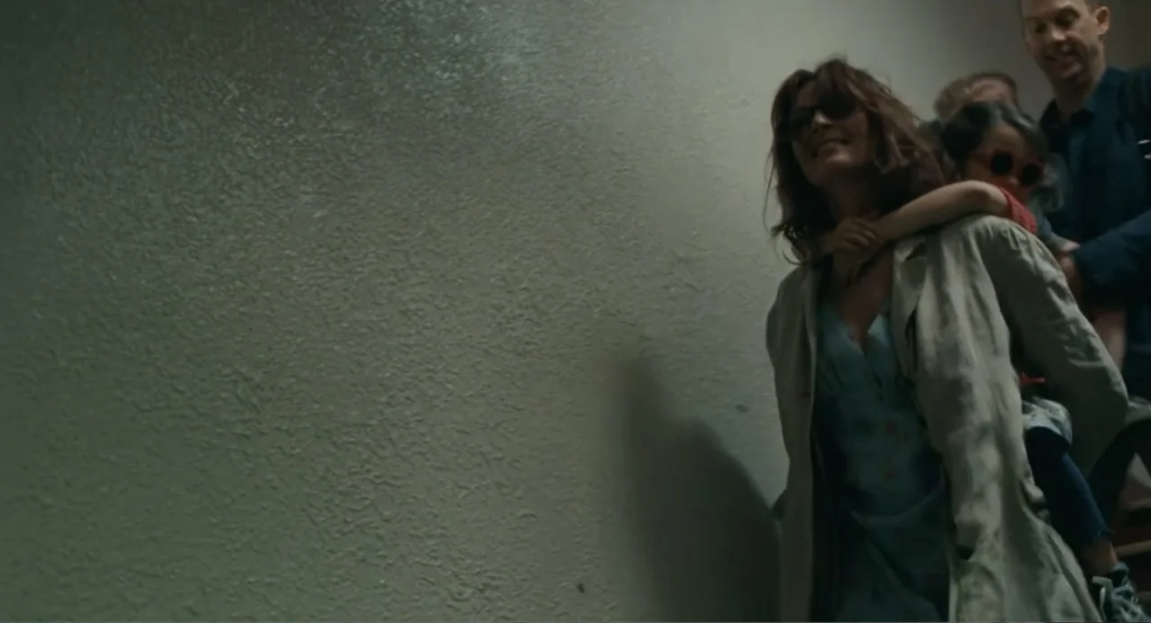 Motherhood (2020) movie still: medium — A dimly lit indoor stairwell with textured off-white walls and concrete surfaces. A woman carries a young…