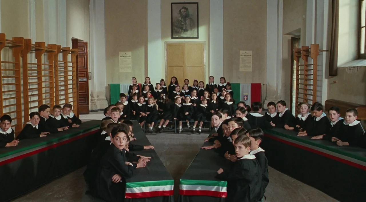 Life is Beautiful (1997) movie still: wide — A high-ceilinged room with gymnastic wall bars and children seated in orderly rows. The students wear…
