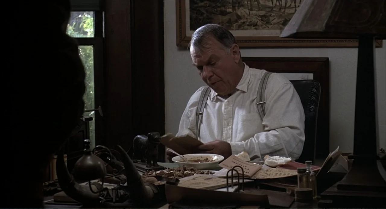 Of Mice and Men (1992) movie still: medium — A cluttered desk is covered with books, papers, a small bowl, and various objects. A man in a white…