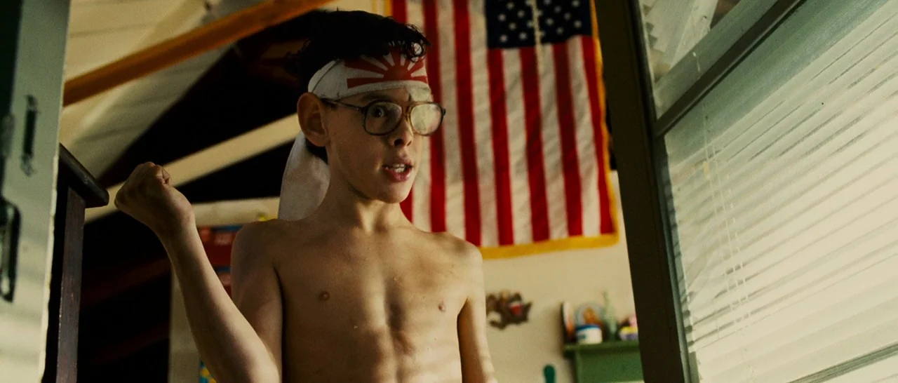 Slice (2018) movie still: medium — An American flag hangs on the wall behind a young boy in a bedroom. The boy wears a headband and glasses while…