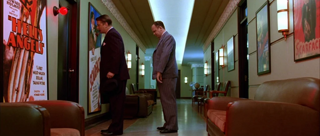 The Aviator (2004) movie still: fullbody — An interior hotel hallway features green walls decorated with framed vintage film posters. Two men in…