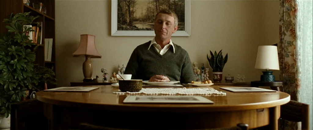 Hunger (2008) movie still: medium — A 1970s dining room features a round wooden table and vintage decorative figurines. A man sits at the table,…