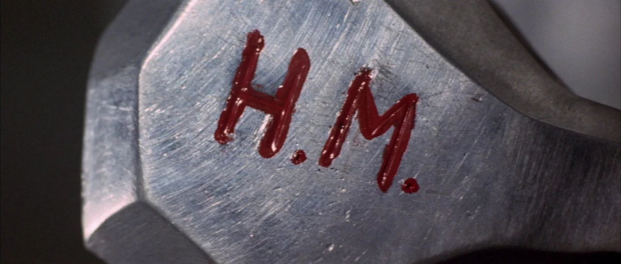 Godzilla Vs Mechagodzilla (1974) movie still: closeup — The surface of a brushed metal object features the hand-painted initials H.M. in red. Thick…