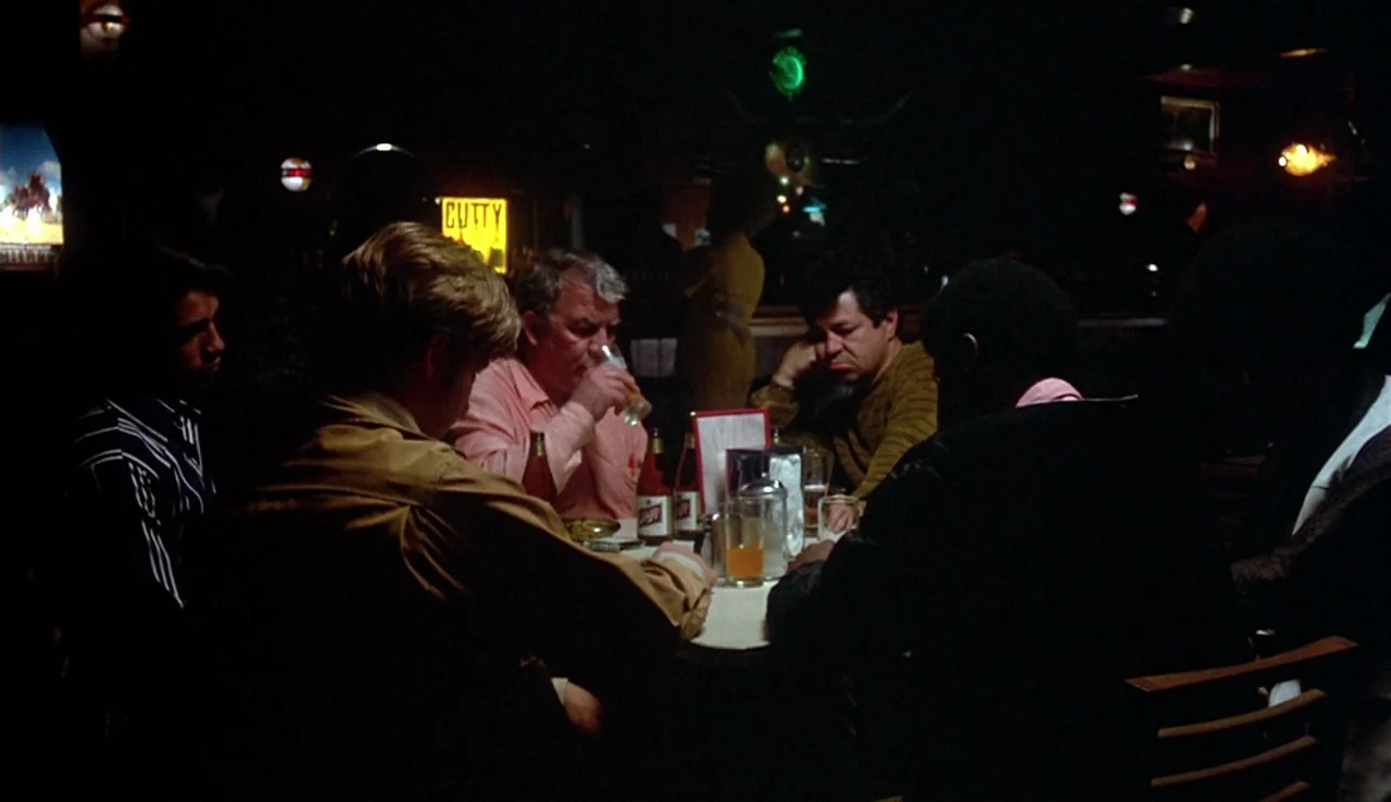 Fat City (1972) movie still: medium — A dark, dimly lit bar with patrons gathered around a small circular table. The table is cluttered with drink…