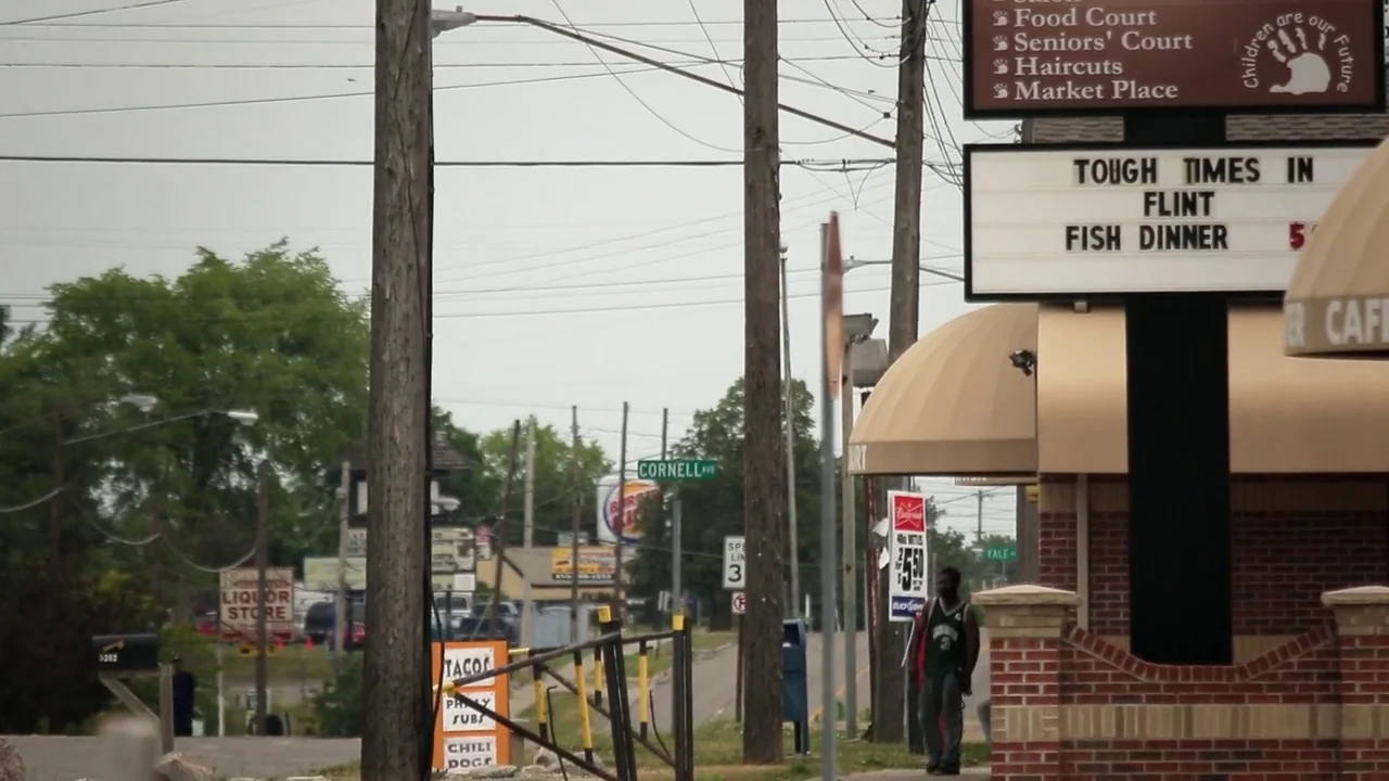 T-Rex (2015) movie still: wide — A suburban roadside with utility poles, road signs, and a brick building exterior. A man walks on the sidewalk near…
