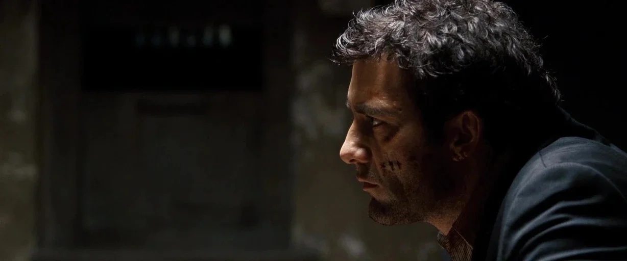 The International (2009) movie still: closeup — A dark, dimly lit room with blurred, indistinct background textures. A man with facial bruising…