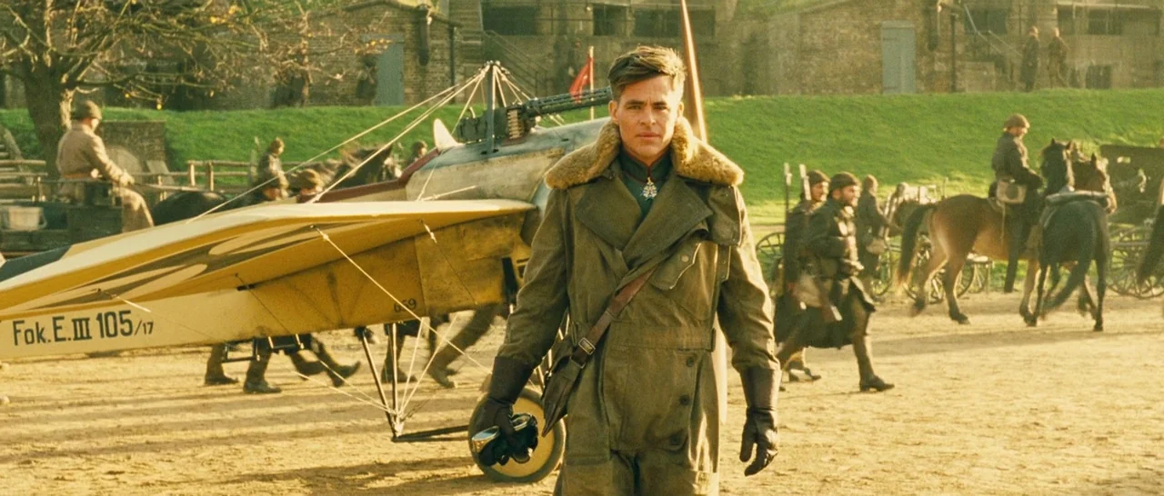 Wonder Woman (2017) movie still: fullbody — A man walks across a dirt field on an active airfield with a biplane. Soldiers and horses populate the…