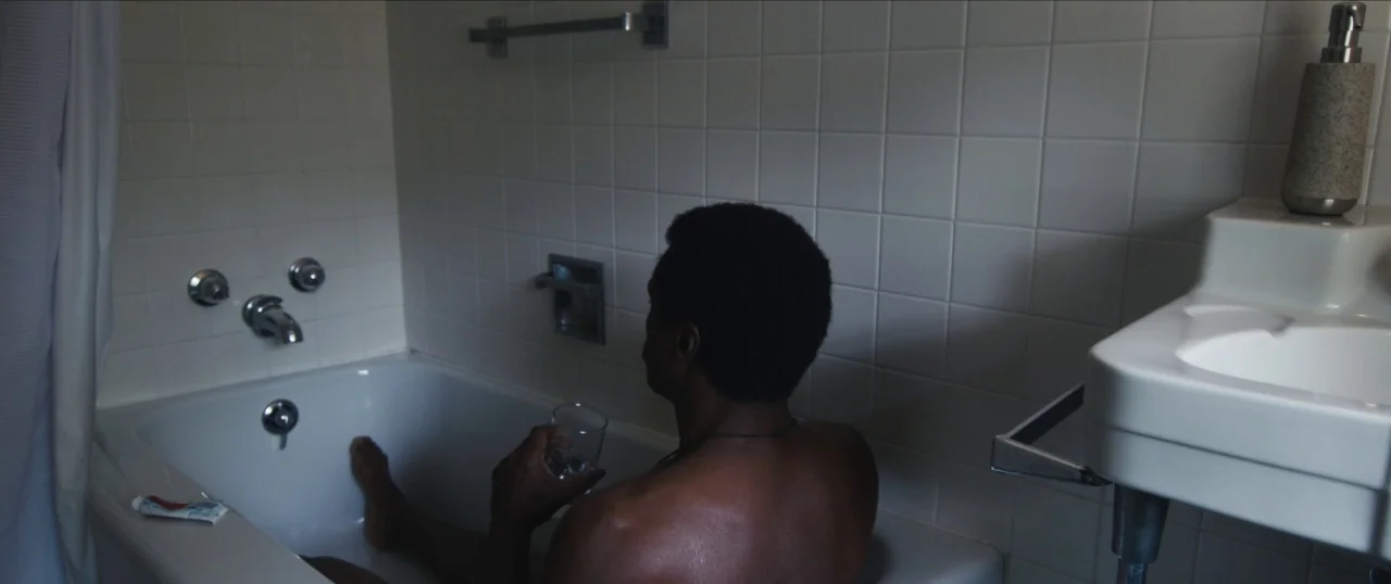 Da Sweet Blood of Jesus (2014) movie still: medium — A white-tiled bathroom features a porcelain bathtub and a wall-mounted sink. A shirtless man…
