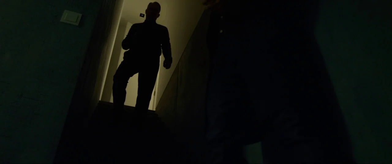 The Girl With The Dragon Tattoo (2011) movie still: medium — A dark, modern interior staircase with deep shadows cast by overhead lighting. One man…