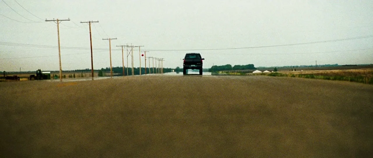 Surveillance (2008) movie still: wide — A dark pickup truck drives away on a flat, wet asphalt road. Wooden utility poles line the shoulder next to…
