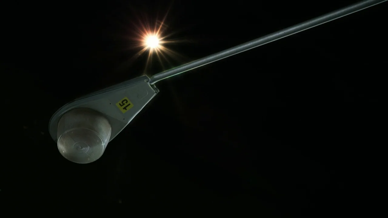 Pass Over (2018) movie still: closeup — A street lamp head with a yellow tag reading 15 against a black sky. The light source creates a visible…