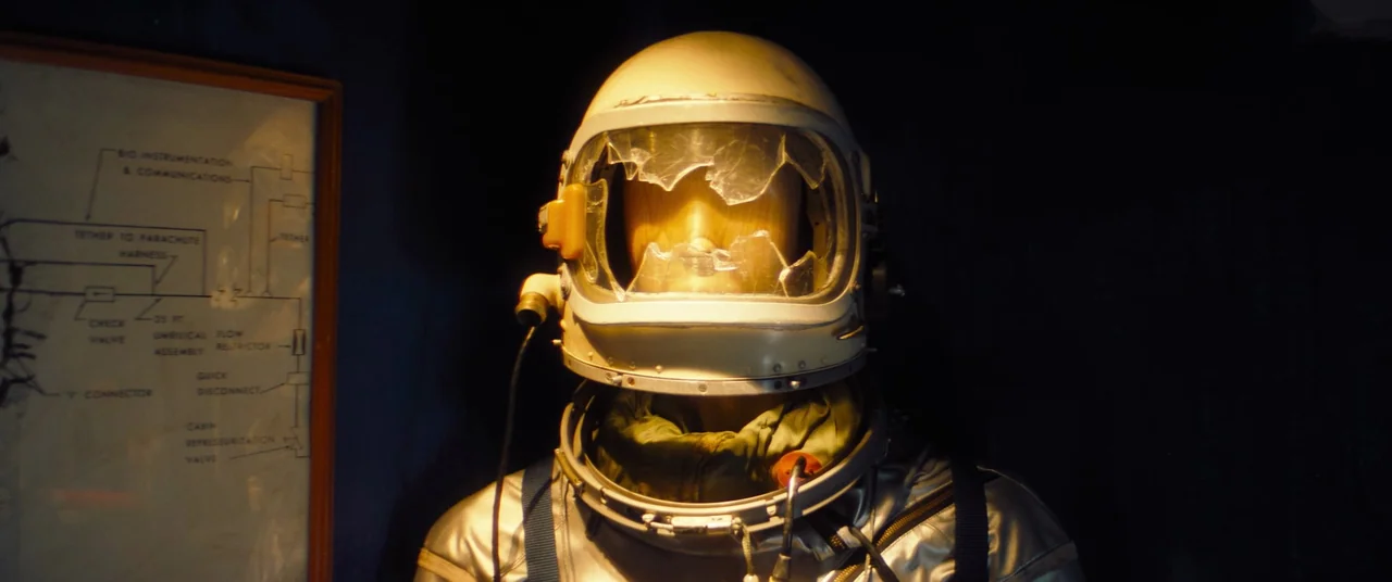 Linoleum (2022) movie still: medium — A vintage silver space suit is displayed in a dimly lit museum setting. A framed technical schematic hangs on…