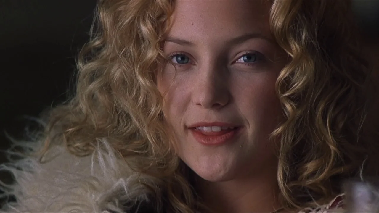 Almost Famous (2000) movie still: closeup — A woman with blonde curly hair looks towards the camera in a dim interior. She wears a textured faux fur…