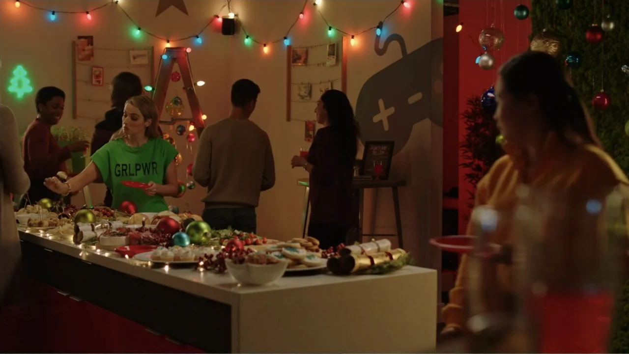 Ghosts of Christmas Past (2021) movie still: medium — A well-lit room decorated for a holiday party features a long buffet table. A young woman in a…