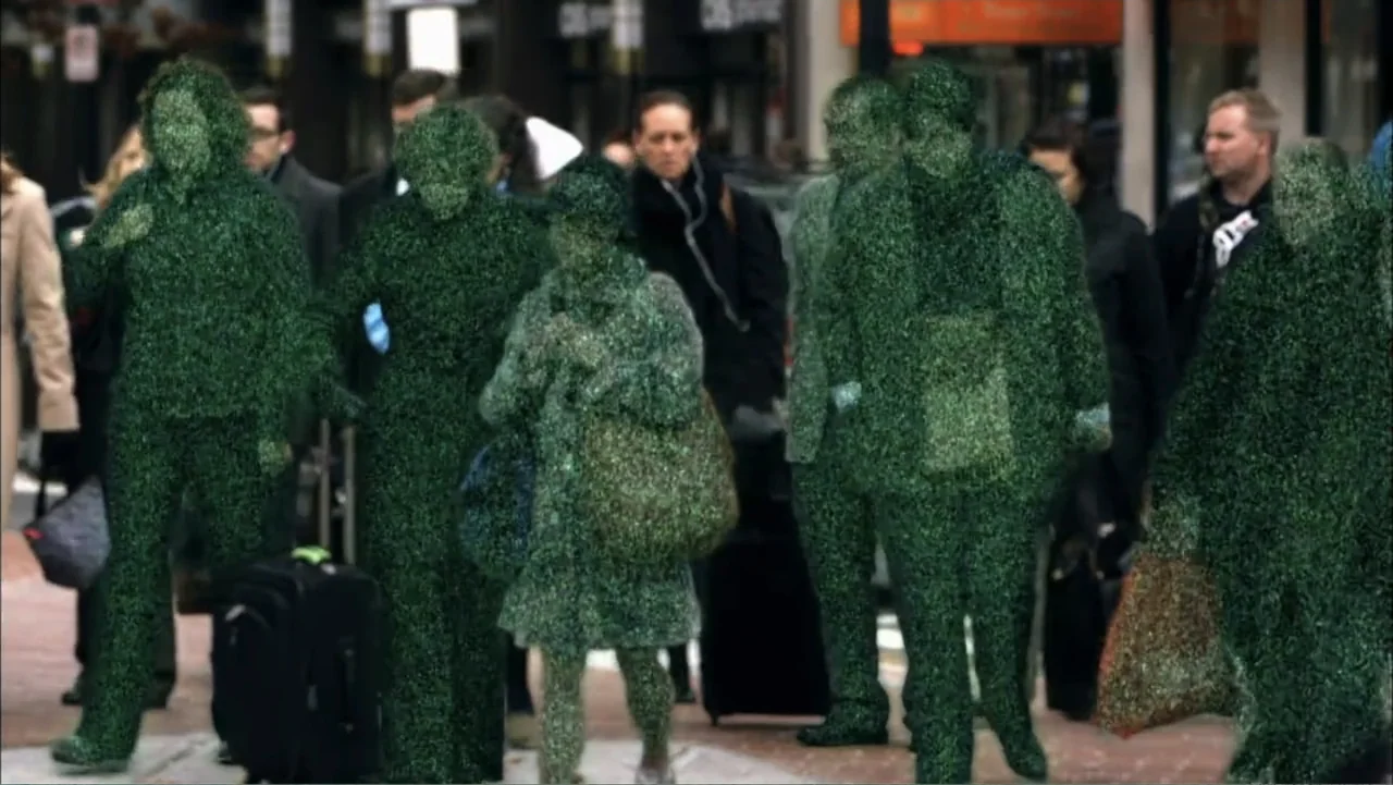 Aliens Inside Us (2013) movie still: medium — A bustling city sidewalk features people wearing suits entirely covered in green synthetic grass.…