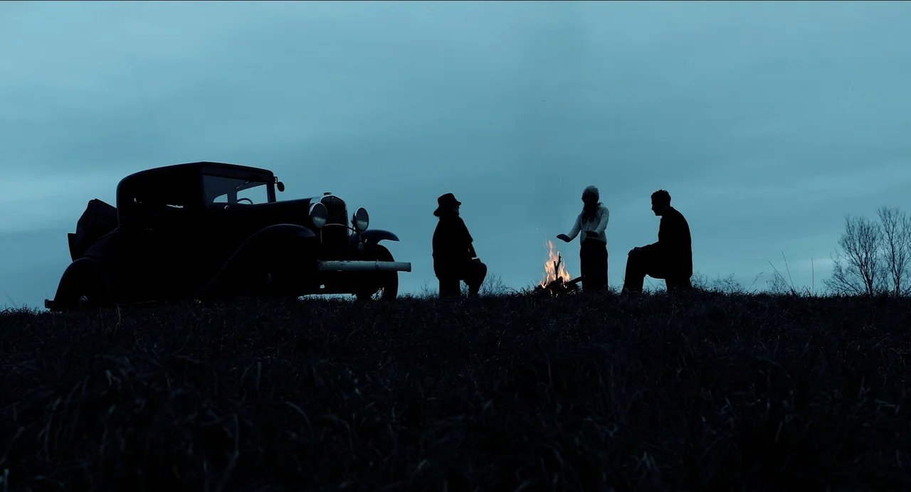 Where The Devil Roams (2023) movie still: wide — Three people huddle around a small fire in a dark field at dusk. A vintage 1930s-style car is…
