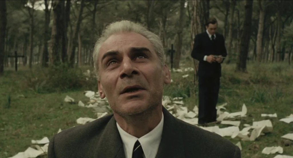 Todo Modo (1976) movie still: closeup — A pine forest clearing is scattered with discarded white papers and wooden grave markers. A man kneels in…