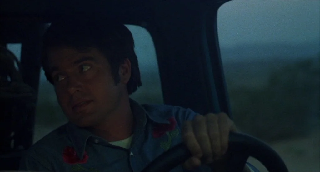 Melvin & Howard (1980) movie still: medium — The interior of a moving vehicle during twilight hours. A man operates the steering wheel while looking…