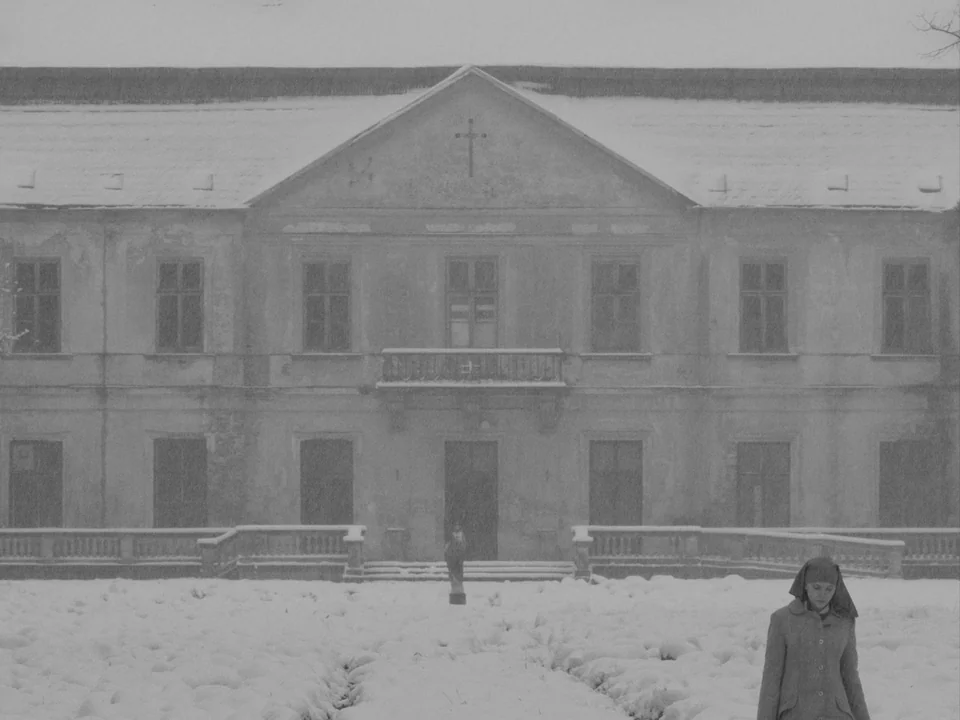Ida (2013) movie still: wide — A stone building with a cross pediment stands behind a snow-covered garden courtyard. A young nun walks across the…