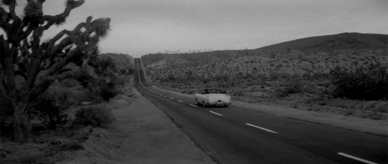 Kiss Me, Stupid (1964) movie still: wide — An asphalt road cuts through a desolate desert landscape with sparse scrub vegetation. A vintage…