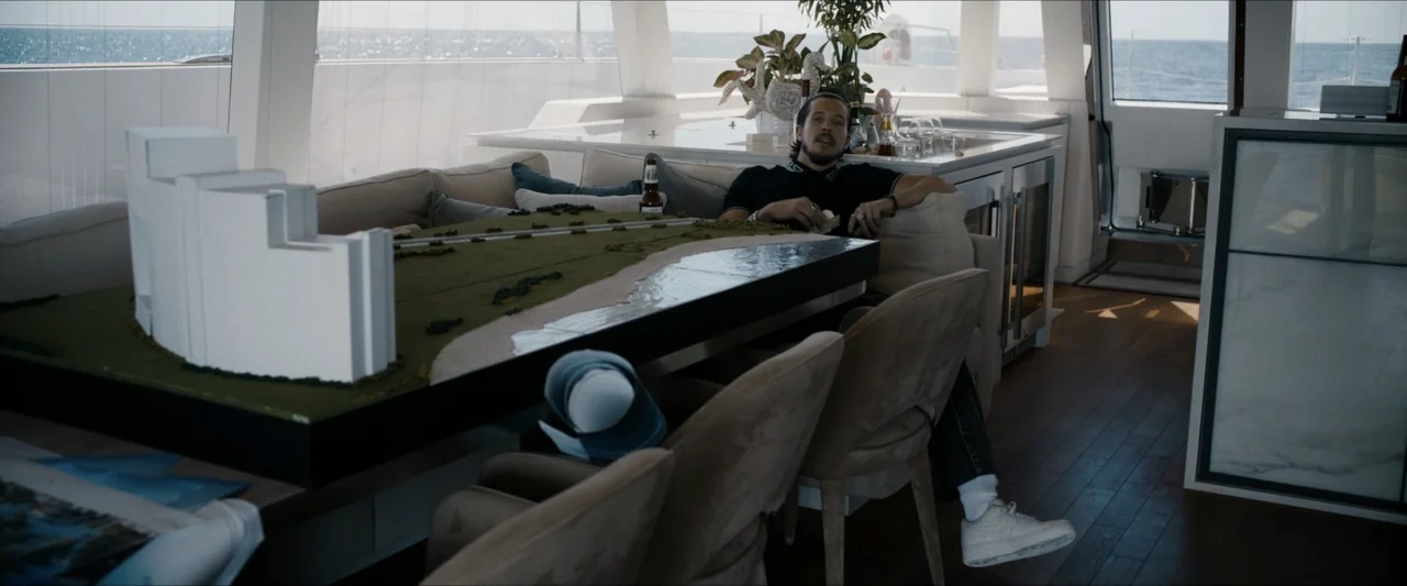 Road House (2024) movie still: medium — A modern yacht interior features a wooden floor, plush seating, and a large table. A man in a dark polo sits…