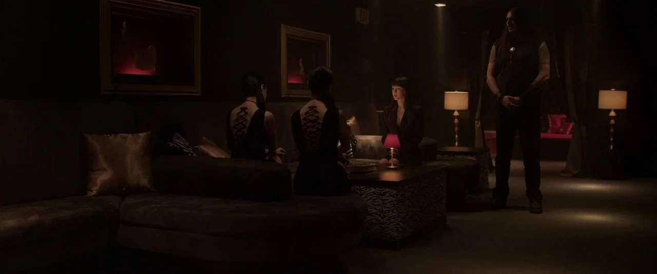 American Mary (2012) movie still: wide — A dark lounge features a large sofa and low tables with amber lamp lighting. Four individuals are gathered…