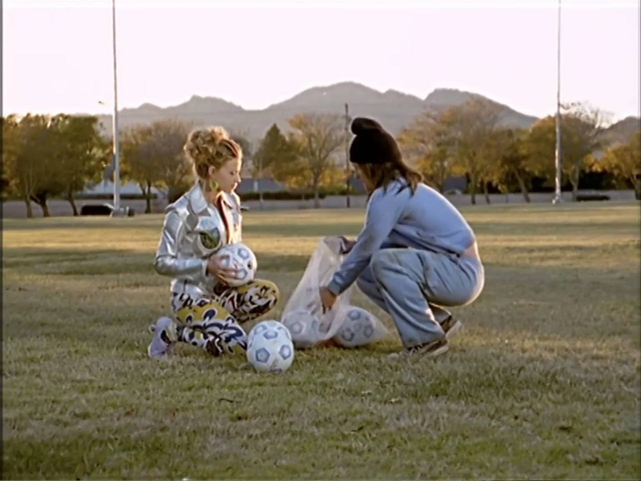 Just 4 Kicks (2003) movie still: fullbody — A flat grassy field at golden hour with distant mountains visible in the background. Two young women…