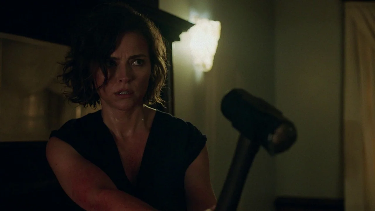 Girl on the Third Floor (2019) movie still: medium — A dimly lit interior space with shadows obscuring the surrounding walls. A woman holds a large…