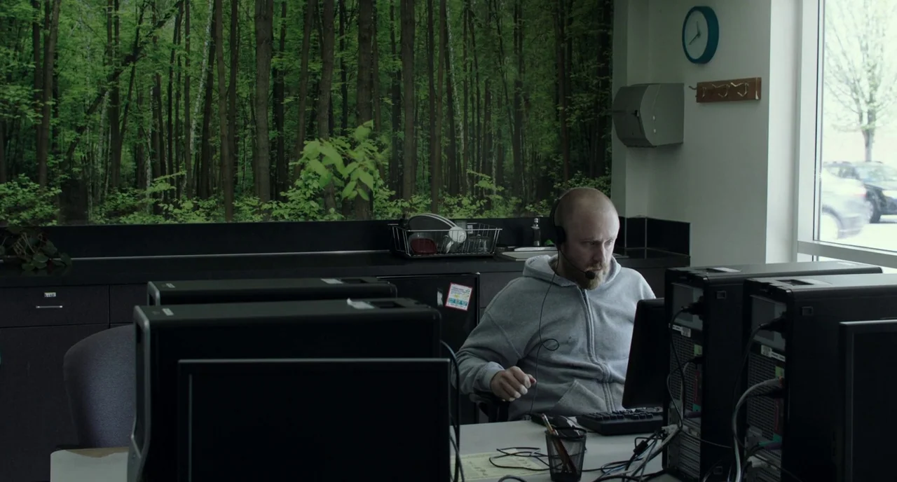 Leave No Trace (2018) movie still: medium — A sterile workspace featuring a large forest-themed wall decal and black desktop computer towers. A man…